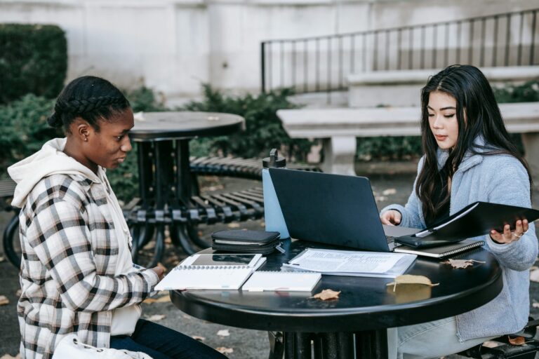Two international students studying together with a laptop and notebooks on a campus patio table